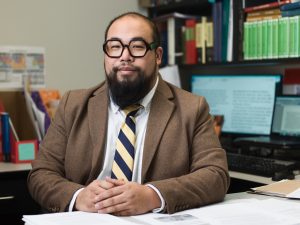 Dr Soon sits at a desk with his hands folded and resting on the desk. He wears large round glasses and blue and yellow stripped tie.
